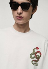 Witte sweatshirt met een grafische afbeelding van een slang in groen en rood, met daaronder de woorden "INTUITIEF STERK BEPALEND". Zwarte zonnebril.