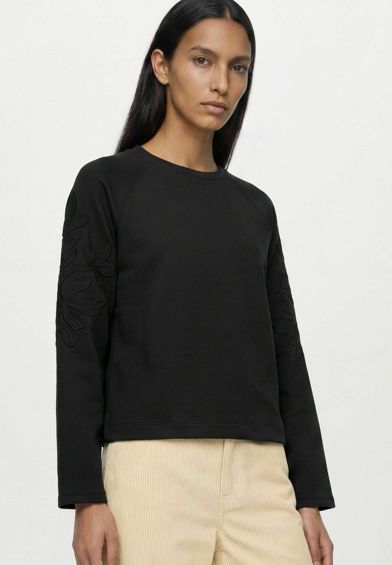 Femme aux longs cheveux foncés portant un sweat-shirt noir avec des broderies florales sur les manches et un pantalon beige clair sur un fond uni.