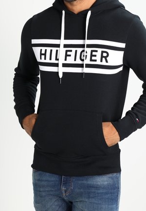 Mężczyzna w czarnej bluzie z kapturem z białym napisem "HILFIGER" i paskami, ręce w przedniej kieszeni, ubrany w niebieskie dżinsy.