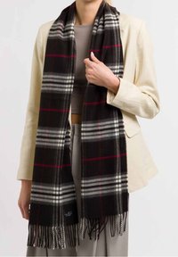 Femme portant un blazer beige et un pantalon gris, tenant une écharpe à carreaux noire avec des rayures blanches et rouges et des bords frangés, sur un haut gris.