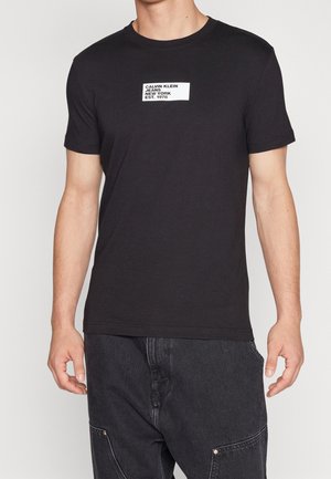 Homme portant un T-shirt noir à manches courtes avec un logo rectangulaire blanc Calvin Klein Jeans New York Est. 1978 sur la poitrine, associé à un jean noir.