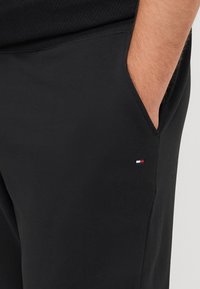 Zwarte sportbroek met een gladde textuur, voorzien van een zijzak en een klein geborduurd logo in rood, wit en blauw.