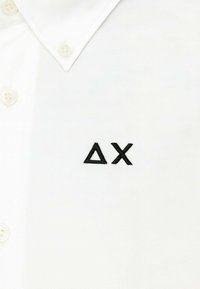 Camicia bianca con bottoni, dalla texture liscia, con un logo ricamato nero formato da un triangolo e una 'X' sul petto.