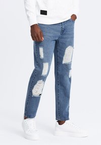 Jeans de mezclilla azul claro con grandes parches desgastados y bordes deshilachados. Llevados con zapatillas blancas y un sudadera blanca.