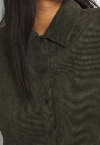 Camicia in corduroy verde oliva con colletto, bottoni frontali e texture a coste verticali con bottoni neri.