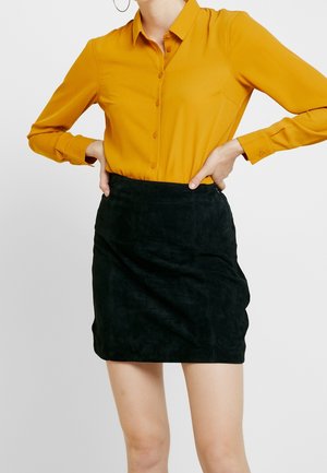 Vrouw draagt een mosterdgele blouse met knopen, ingestopt in een zwarte suède minirok, met handen in de zij.