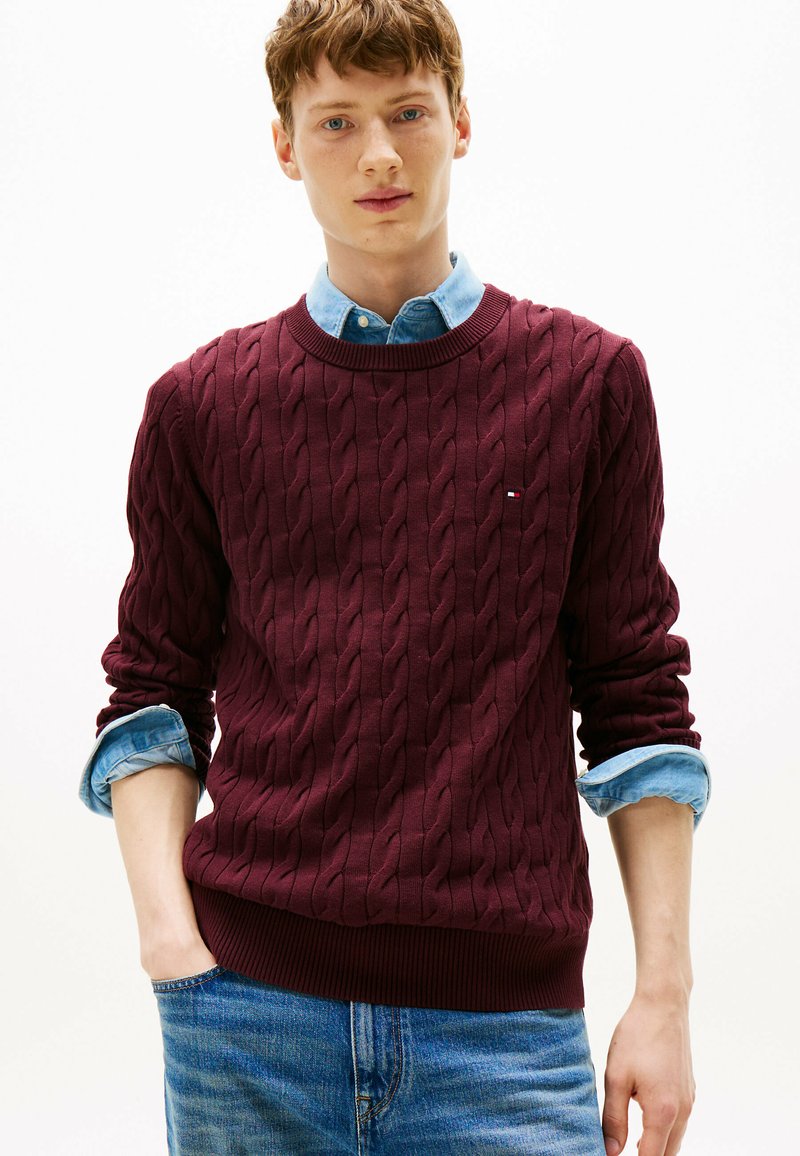 Burgundy cable-knit sweater with ribbed hem, worn over a light chambray shirt, paired with light blue jeans.