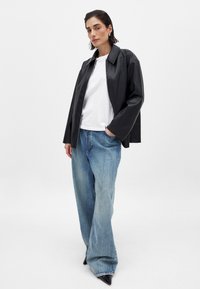 Giacca nera in similpelle con colletto a punta, indossata sopra una t-shirt bianca, abbinata a jeans larghi di un blu chiaro e tacchi a punta neri.