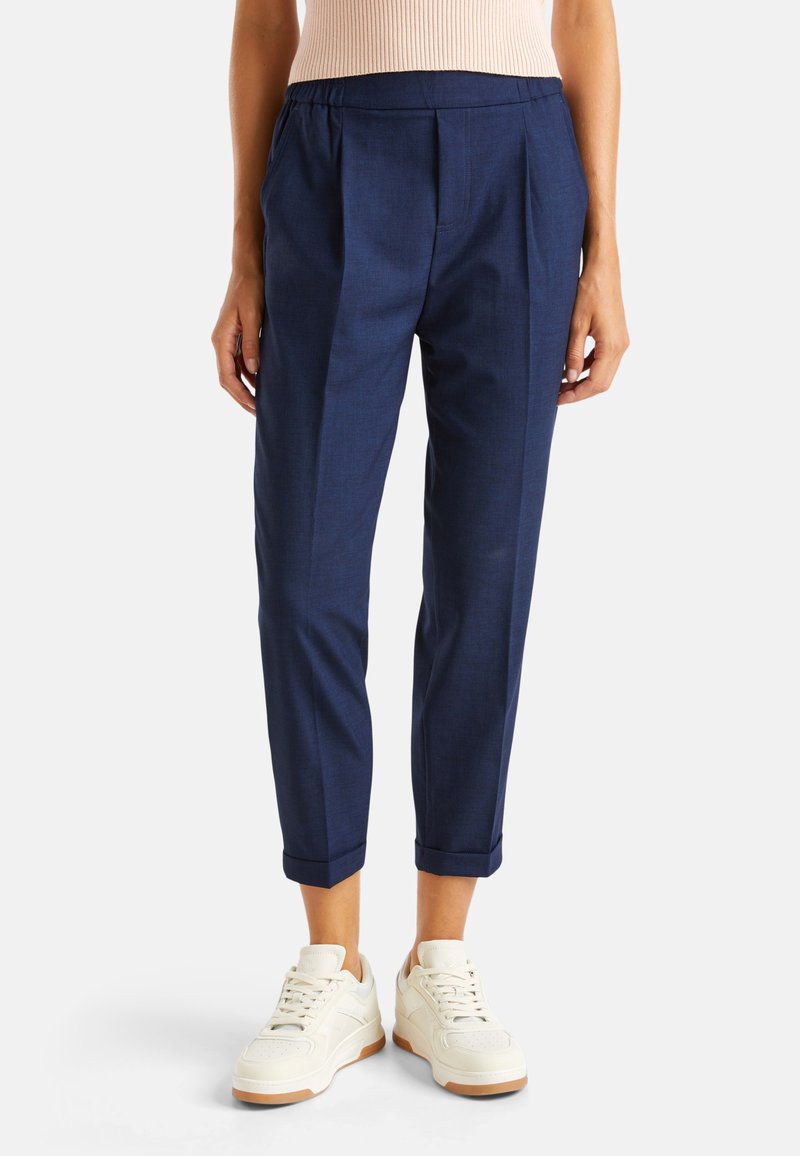 Granatowe spodnie o dopasowanym kroju z plisowanym przodem, elastycznym paskiem w talii i podwiniętymi mankietami, zestawione z białymi trampkami. Gładka faktura.