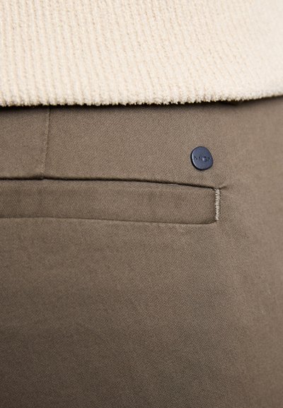 Olivengrønne bukser med en glat tekstur og en baglomme. Inkluderer en cirkulær mørkeblå logo-knap, delvist dækket af en cremefarvet sweater.