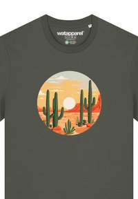 Groene katoenen t-shirt met een cirkelvormig woestijnlandschap met cacti en een zonsondergang, weergegeven in warme kleuren en een grafische ontwerpstijl.