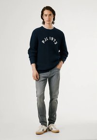 Marineblå ribbestrikket genser med hvit tekst "PJL 1973", kombinert med lysegrå jeans og hvite joggesko med brune detaljer. Modellen står med en hånd i lommen.