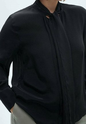 Vrouw die een zwarte blouse met lange mouwen en een gestrikt halsdetail draagt, en lichtgrijze broek, hand in zak, tegen een effen achtergrond.