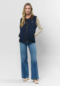 Námornícky prešívaný vest s hnedým golierom, cvokmi a bočnými vreckami, nosený cez pruhované tričko s dlhým rukávom a v kombinácii so širokými džínami.