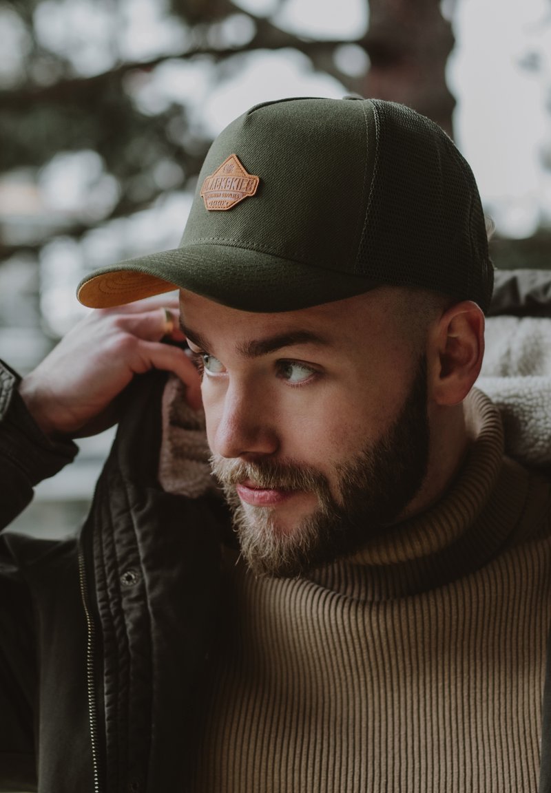 Cappello verde con una toppa in pelle beige, visiera curva e pannelli laterali in rete. Il modello indossa un dolcevita a coste marrone e una giacca nera.