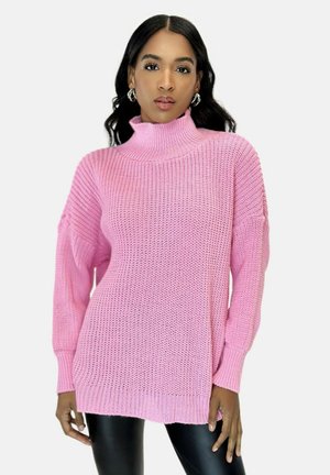 Różowy sweter z dzianiny z wysokim golfem, teksturowanym wykończeniem, oversize'owymi rękawami i luźnym fasonem. Noszony z czarnymi legginsami.
