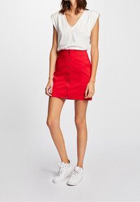 Frau in ärmelloser weißer V-Ausschnitt-Bluse, knopfbarer roter Minirock und weißen High-Top-Sneakern, stehend vor einem hellen Hintergrund.