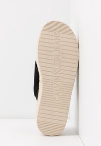 Semelle d'une chaussure Calvin Klein Jeans beige avec des lignes d'adhérence horizontales, reposant verticalement contre un mur d'angle blanc.