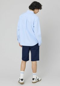 Chemise boutonnée bleu clair à manches longues, associée à un short bleu marine. Baskets blanches avec des accents noirs et des chaussettes rayées. Tenue décontractée.