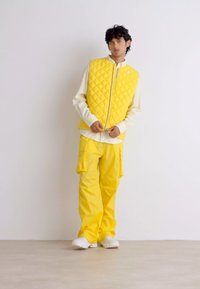 Gilet matelassé jaune avec fermeture éclair, chemise à manches longues blanche et pantalon cargo large jaune. Le mannequin porte des baskets blanches et se tient sur un sol beige.