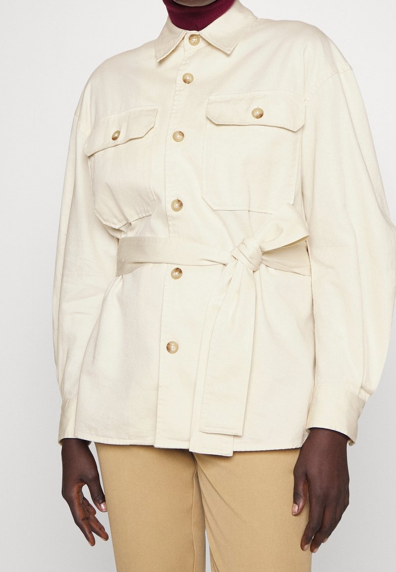 Kremowa zapinana na guziki kurtka z klapkowymi kieszeniami i paskiem w talii, noszona przez osobę o ciemnej karnacji, z widocznymi rękami, na tle beżowych spodni.