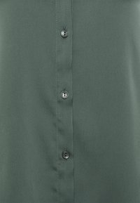 Green button-up shirt made of smooth fabric, featuring a vertical row of black buttons and subtle stitching detail.