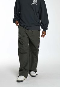 Olijfgroene wijde broek met geborduurde ontwerpen op de bovenbenen, gecombineerd met een donkere sweatshirt en witte schoenen.