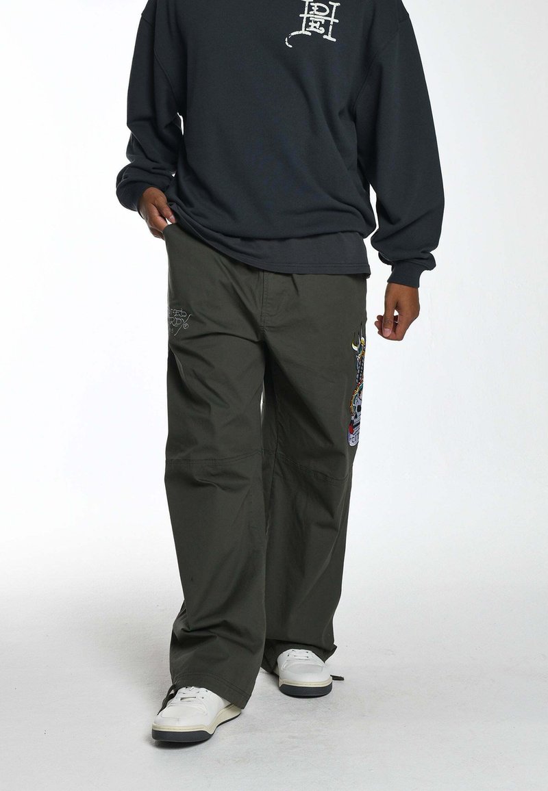 Olijfgroene wijde broek met geborduurde ontwerpen op de bovenbenen, gecombineerd met een donkere sweatshirt en witte schoenen.