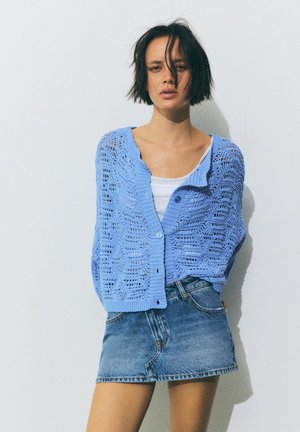 Rövid sötét hajú nő, világoskék kötött kardigánt visel fehér felső és kék farmer miniszoknya fölött, egy sima falnak támaszkodva áll.