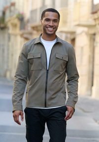 Khakifarbene Zip-Jacke mit zwei Brusttaschen, getragen über einem weißen T-Shirt, kombiniert mit dunklen Hosen; glatte Textur und taillierte Passform.