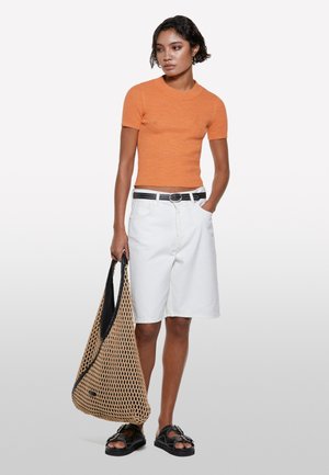 Top arancione a righe con maniche corte, pantaloni corti bianchi fino al ginocchio, cintura nera, sandali neri e una borsa tessuta color beige con fodera nera.