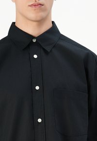 Close-up van een persoon die een zwart overhemd met knopen en een kraag draagt, met een borstzakje, met de focus op de bovenste torso en nek.