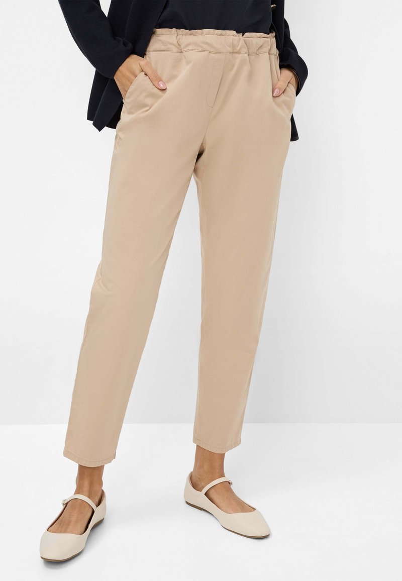 Beige, lichte, taps toelopende broeken met een elastische tailleband en zijzakken. Gladde textuur; gecombineerd met beige platte schoenen.