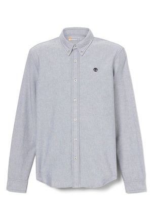 Mężczyzna w jasnoszarej koszuli z guzikami na białym graficznym t-shircie, ciemnoniebieskich spodniach jeansowych podwiniętych u dołów, brązowym pasku i beżowych butach roboczych stojący.