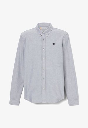 Homme portant une chemise à boutons gris clair sur un t-shirt graphique blanc, un jean bleu foncé avec les ourlets roulés, une ceinture marron et des bottes de travail beige, debout.