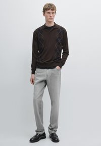 Maglione marrone a maniche lunghe con motivo a diamante, abbinato a jeans in denim grigio chiaro e scarpe in pelle nere. Colletto piatto e polsini a coste.