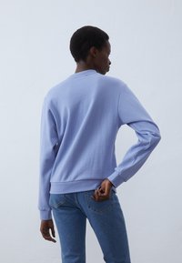 Felpa lilac con maniche lunghe, polsini e orlo a coste; indossata con jeans blu. Tessuto morbido con una vestibilità casual, vista da dietro.