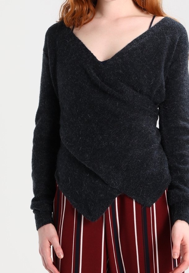 Femme portant un pull noir en style cache-cœur en tissu doux avec un col en V profond et un pantalon large à rayures rouges, debout devant un fond uni.