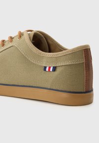 Olive green canvas sneaker with tan laces, brown leather accents, rubber sole, and a small tri-color logo patch near the heel.