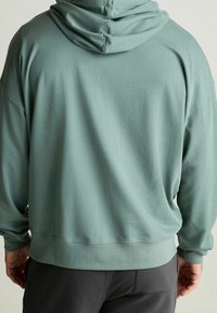 Světle zelená mikina vyrobená z měkké tkaniny. Má velkou, dvakrát podloženou kapuci, kleslé ramena a žebrovaný lem a manžety.
