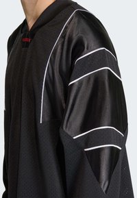 Crna atletska majica s teksturiranim mrežastim tijelom, glatkim rukavima od tkanine s bijelim prugama i kapuljačom na izrezu oko vrata.