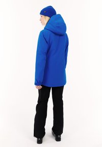 Chaqueta impermeable azul con capucha, puños ajustables y un corte relajado, combinada con pantalones de esquí negros y botas negras.