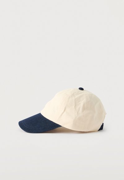 Casquette de baseball en coton beige avec une visière en denim bleu foncé. Dispose d'une couronne arrondie et d'un petit bouton sur le dessus.