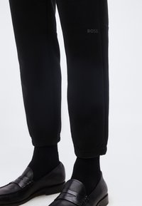 Μαύρο στενό παντελόνι με μανσέτες στα άκρα και λογότυπο "BOSS" στον μηρό, φορεμένο με μαύρες κάλτσες και μαύρα δερμάτινα μοκασίνια σε λευκό φόντο.