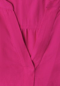 Tissu rose vif avec encolure en V et détail de couture froncée sous la zone du col, montrant texture et coutures.