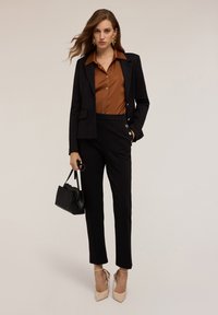 Blazer noir ajusté et pantalon, associés à une chemise en coton marron à boutons. Accessoirisé avec un sac à main noir et des chaussures à talons blocs beiges.
