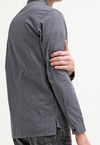 Camisa gris de manga larga con cuello y puños abotonados, de textura suave. Presenta un dobladillo recto y detalles de costura sutiles.