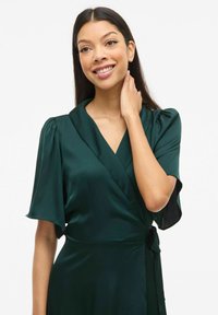 Smaragdgrønn sateng omslagskjole med korte puffermede ermer, V-hals og detalj i midjebånd. Glatt tekstur, elegant design og flatterende silhuett.