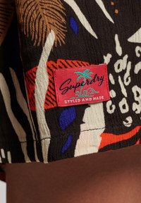 Tela estampada colorida con una etiqueta roja "Superdry Styled and Made" que presenta palmeras en color teal en el borde inferior de una prenda.
