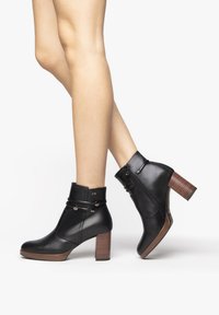 Stivali alla caviglia in pelle nera con tacco grosso, doppi cinturini con accessori in argento, chiusura con zip laterale e tacco in legno texturizzato.